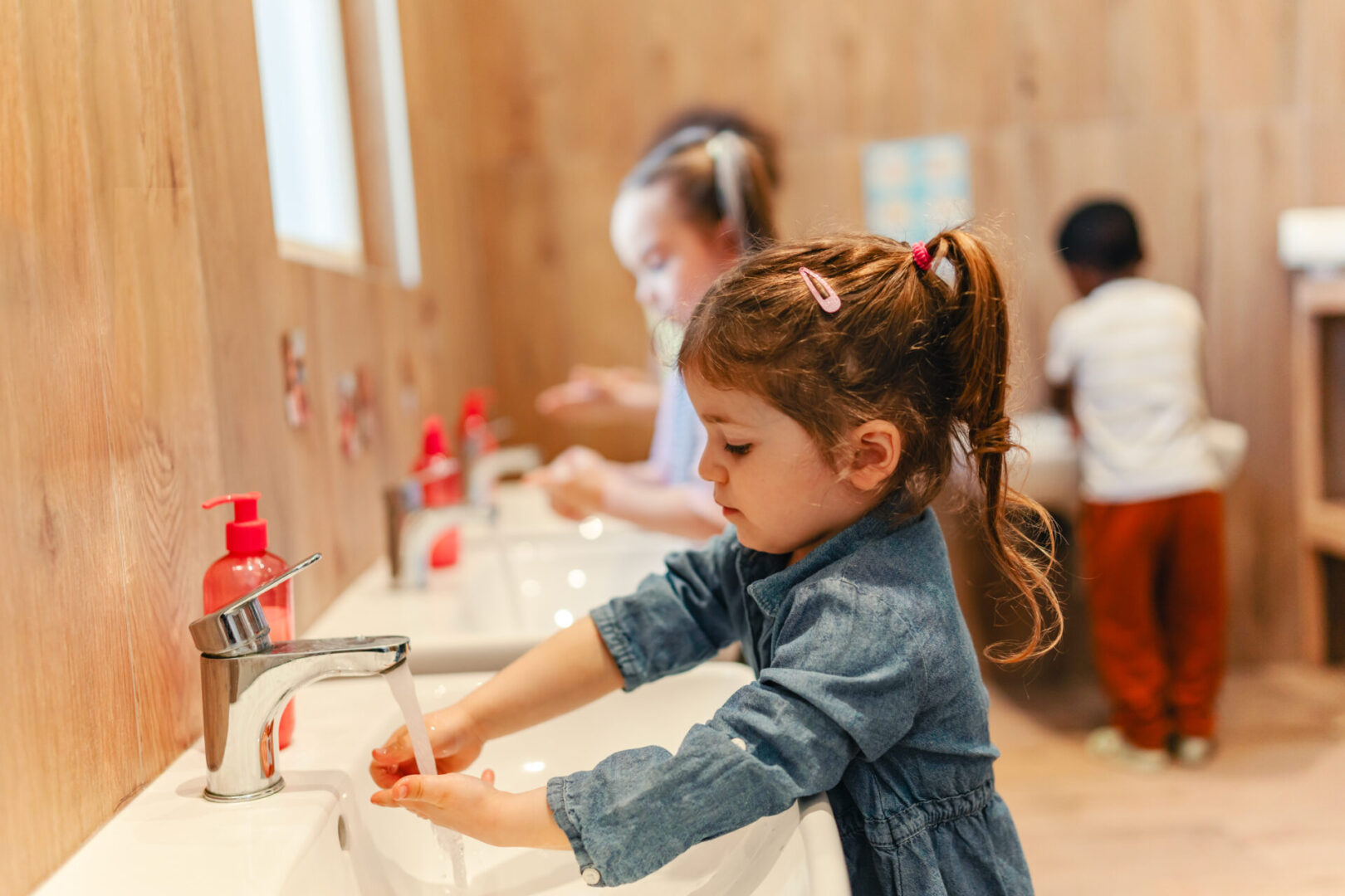 Multicultural preschool children wash their hands at sinks in a wooden-themed bathroom, practicing hygiene routines as part of their daily classroom activities. Calm Kids: Anxiety Skills Center